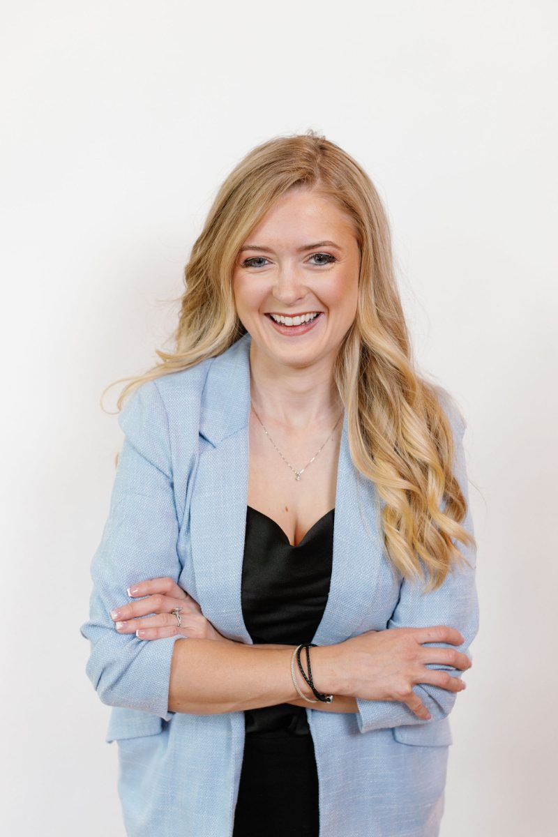 Smiling Catskills wedding planner in a light blue blazer poses against a white wall, a warm first impression for couples hiring a wedding planner.