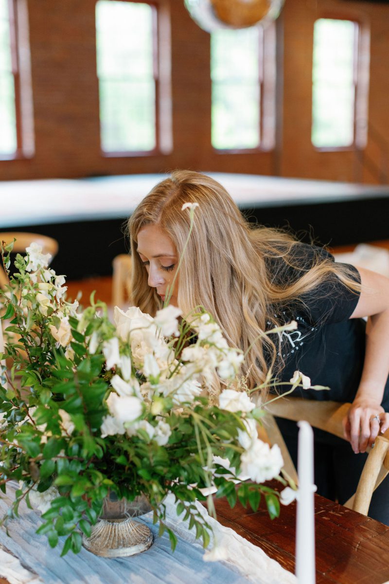 Catskills wedding planner leans in to smell a white floral centerpiece on a reception table, checking each detail so couples hiring a wedding planner can relax on the day.