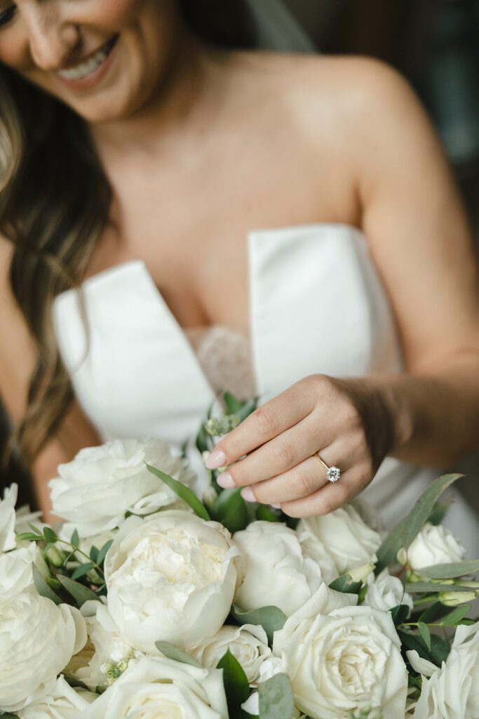 Bride smiles while touching her white rose bouquet, showing off engagement ring.