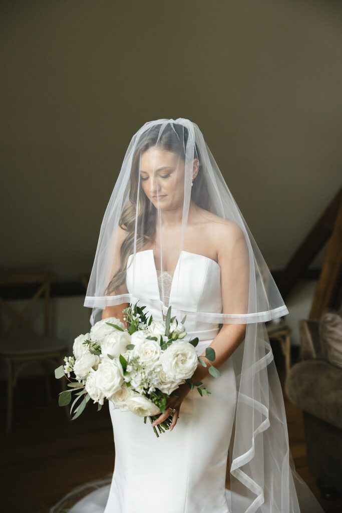 Bride in strapless gown with veil, holding a white rose bouquet, standing gracefully before the ceremony.