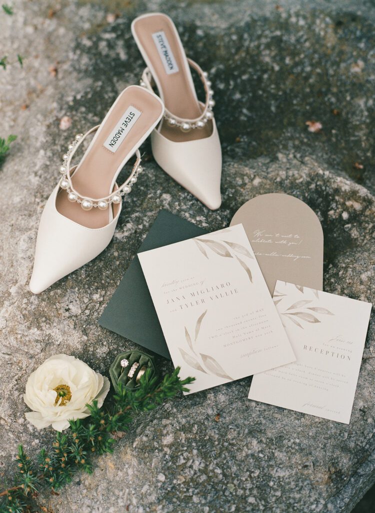 flat lay green and beige wedding stationery and bridal shoes with wedding rings and white flower.