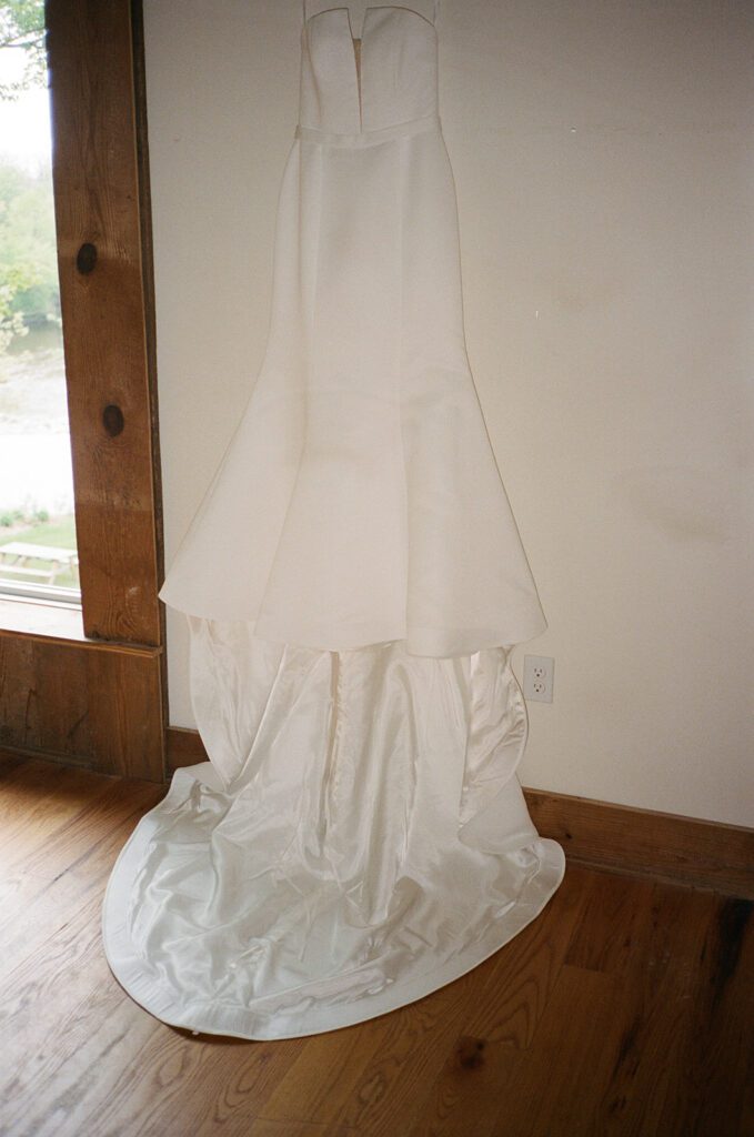 Bride’s strapless fit-and-flare wedding gown hanging by the window before ceremony.
