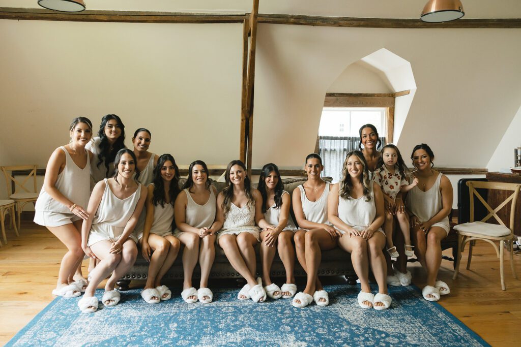Bride and bridesmaids in matching loungewear slippers sitting together on sofa, laughing.