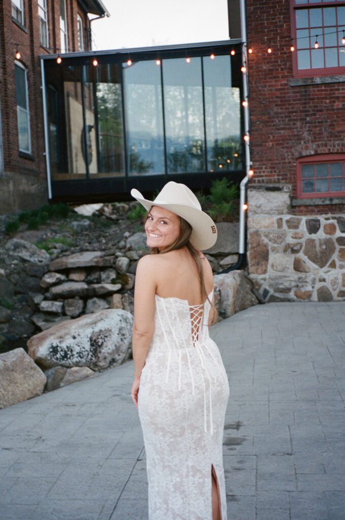 Bride in lace gown and cowboy hat smiles back at the camera while standing outside the brick winery, glowing under string lights.