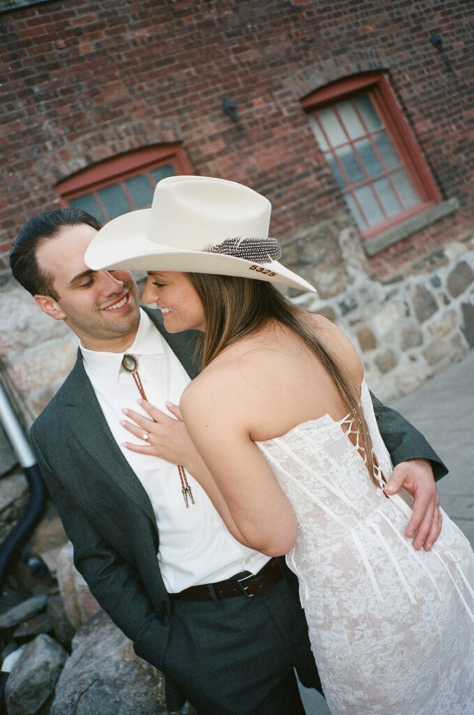 Bride and groom share a joyful moment outside, her hand on his chest showing her ring, with rustic brick walls behind them.