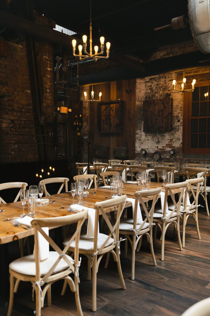 Warmly lit dining tables at City Winery with rustic wood details, set with glasses, napkins, and silverware for the celebration.