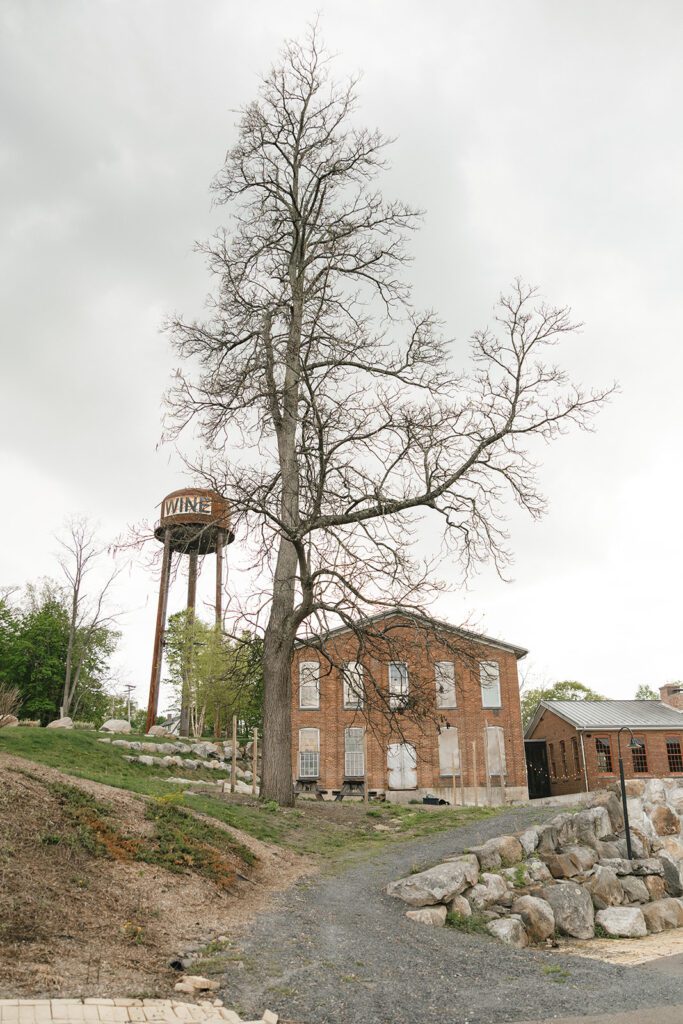 Scenic exterior view of City Winery, with a historic brick building, tall water tower, and trees along a rustic gravel path.