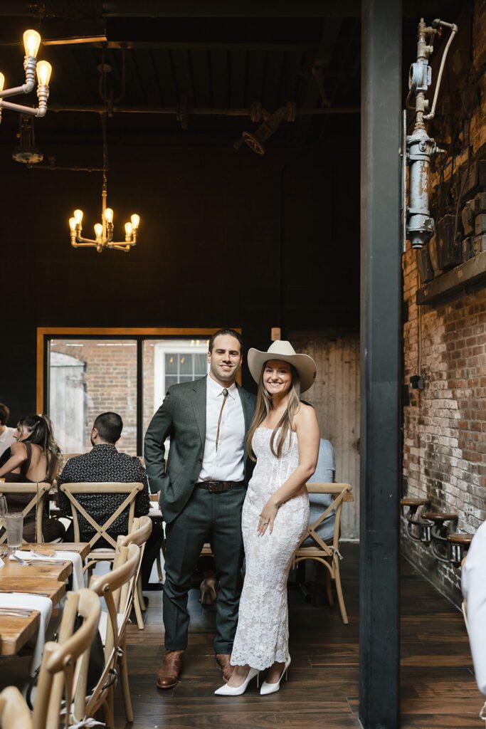 Bride and groom stand together at City Winery, she in a lace dress and cowboy hat, he in a suit, with rustic brick and wood décor in the background.