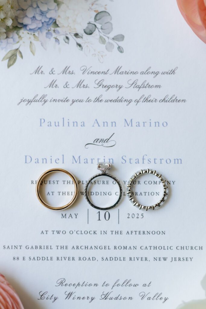 Close-up of wedding invitation with floral design and three rings displayed, including gold band, diamond ring, and eternity band.