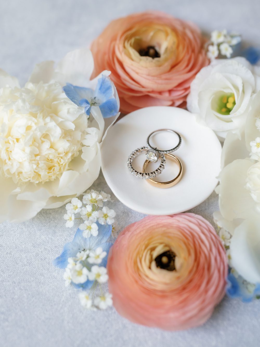 White and blush blooms with blue accents and wedding rings sitting on top of a small white dish.