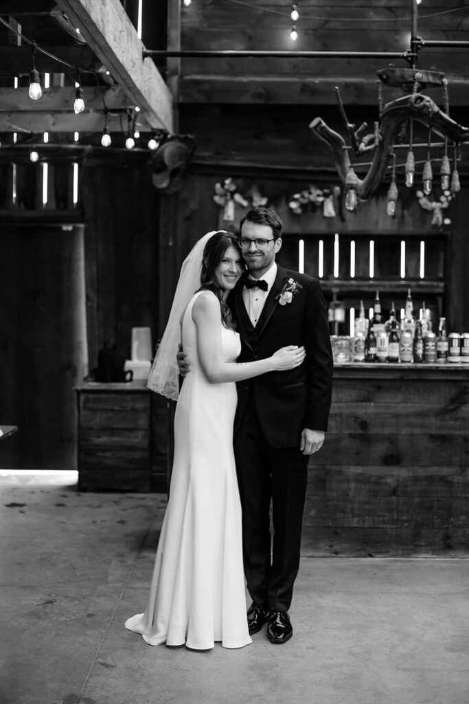 Bride and groom embrace and smile in front of a rustic barn bar at The Arnold House.