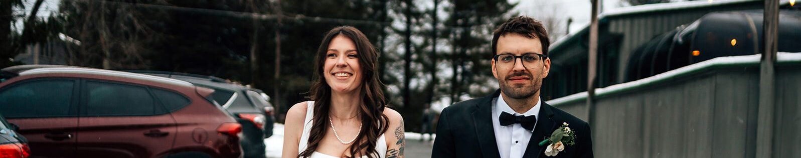 A bride in a white dress with tattoos and a pearl necklace walks hand-in-hand with the groom in a black tuxedo, smiling together on a snowy road with cars and trees in the background.