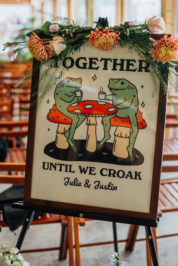 Wedding sign with two cartoon frogs on mushrooms reads “Together Until We Croak.”