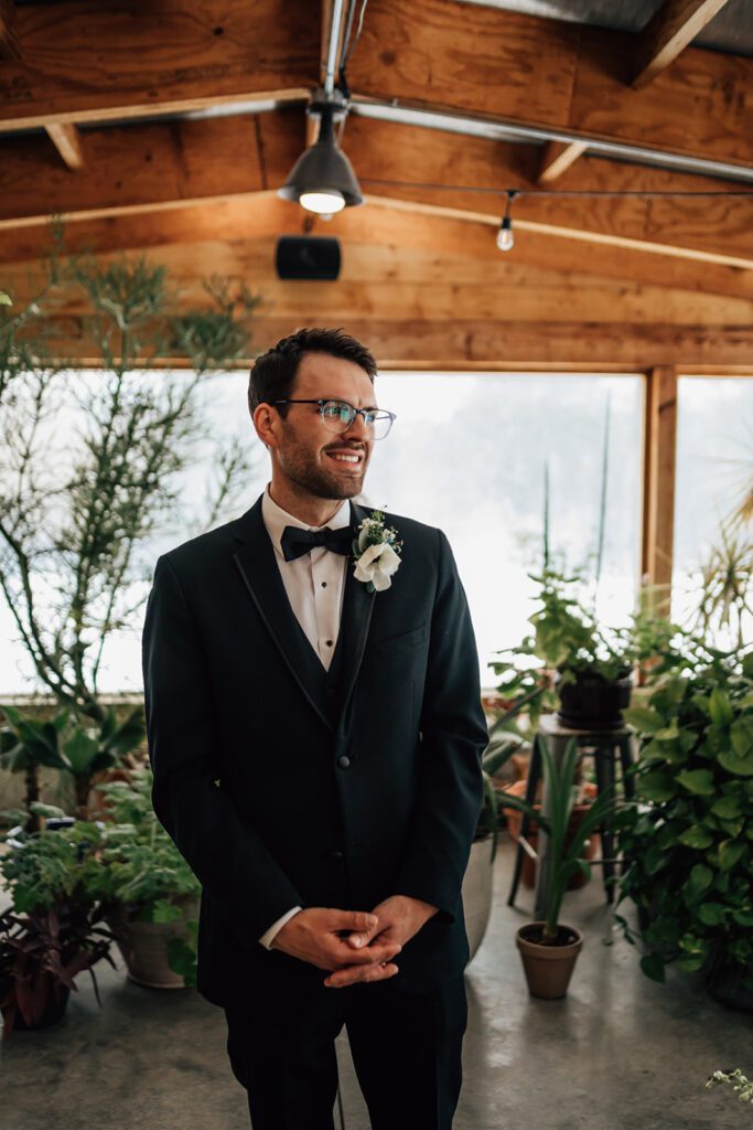 Groom smiles in a black tuxedo with boutonniere, standing among plants at The Arnold House.