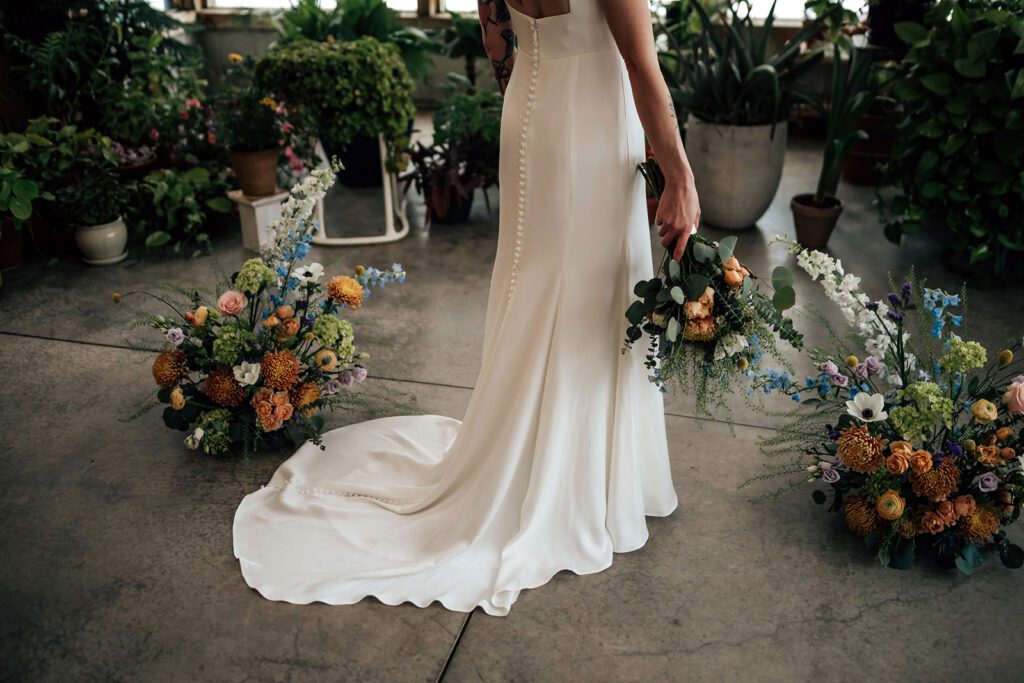 Bride in satin gown holds bouquet among colorful floral arrangements.