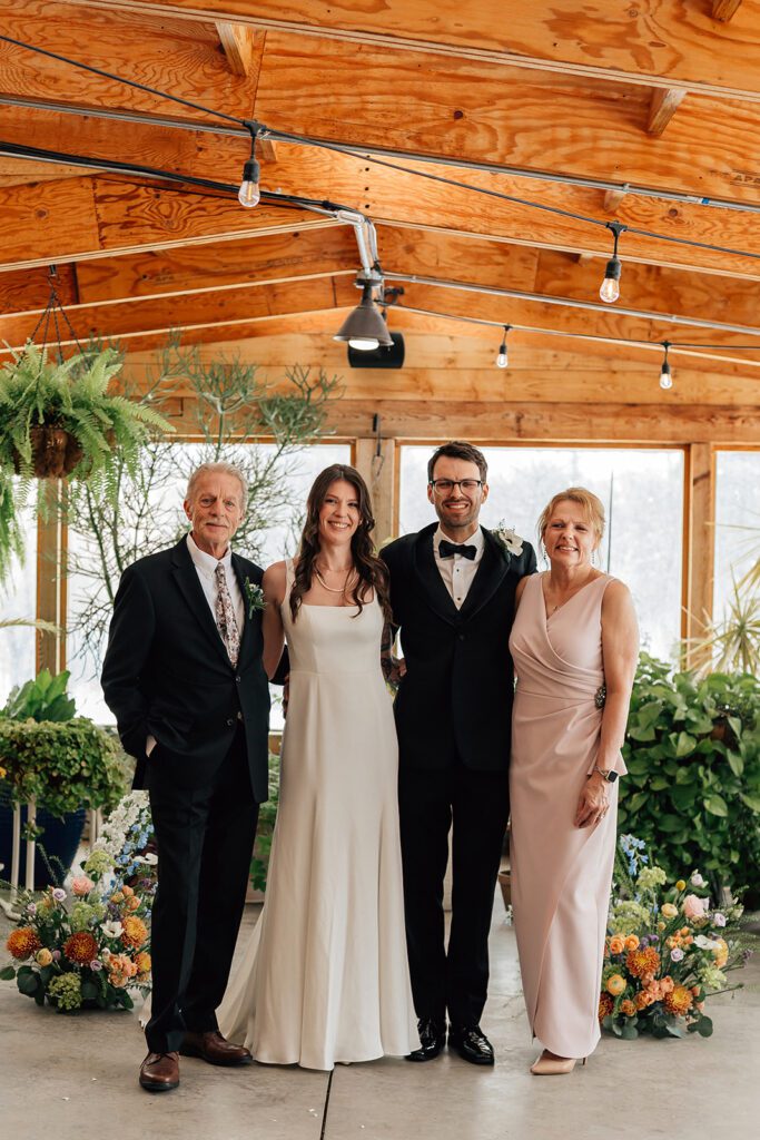 Bride and groom pose with parents, smiling inside The Arnold House greenhouse space.