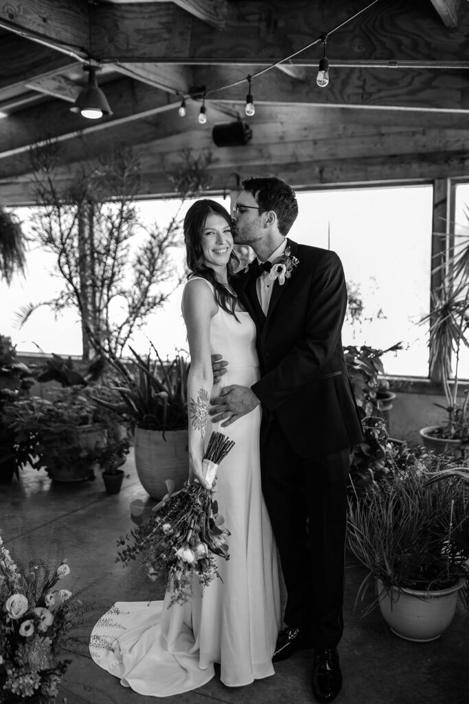 Groom kisses bride’s forehead as she smiles, bouquet in hand, surrounded by greenery at The Arnold House.