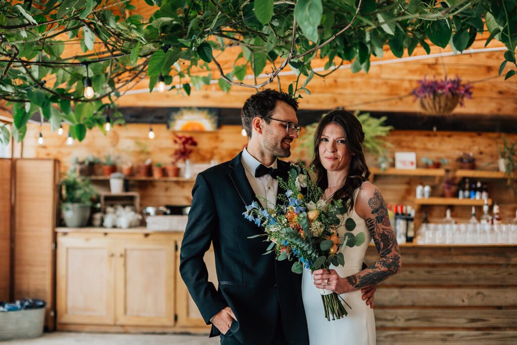 Groom in a black tuxedo smiles at the bride holding a colorful bouquet under greenery at The Arnold House.