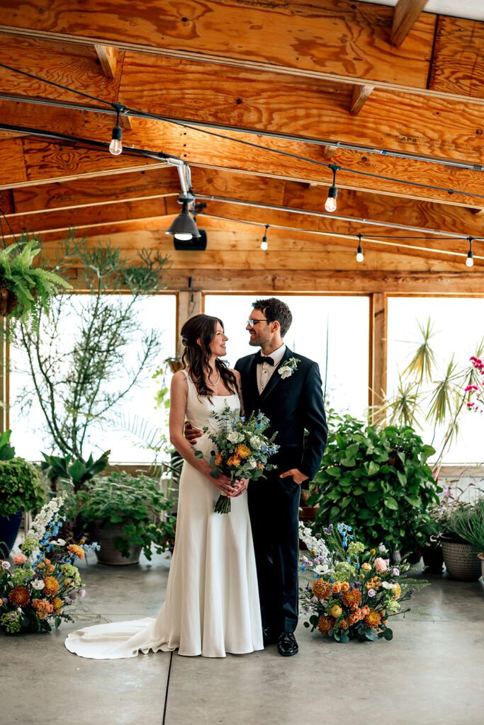 Bride and groom stand facing each other, smiling with floral arrangements around them at The Arnold House.