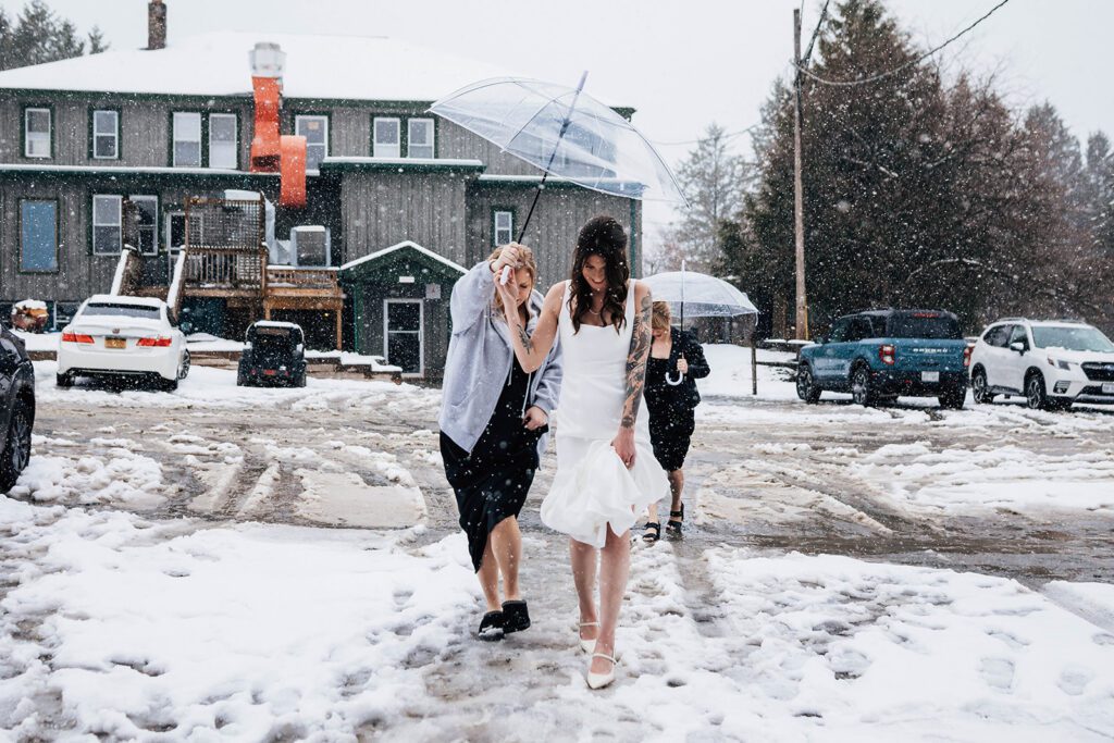 Bride walks through snow at The Arnold House with friends holding umbrellas, her white dress lifted from the slush.
