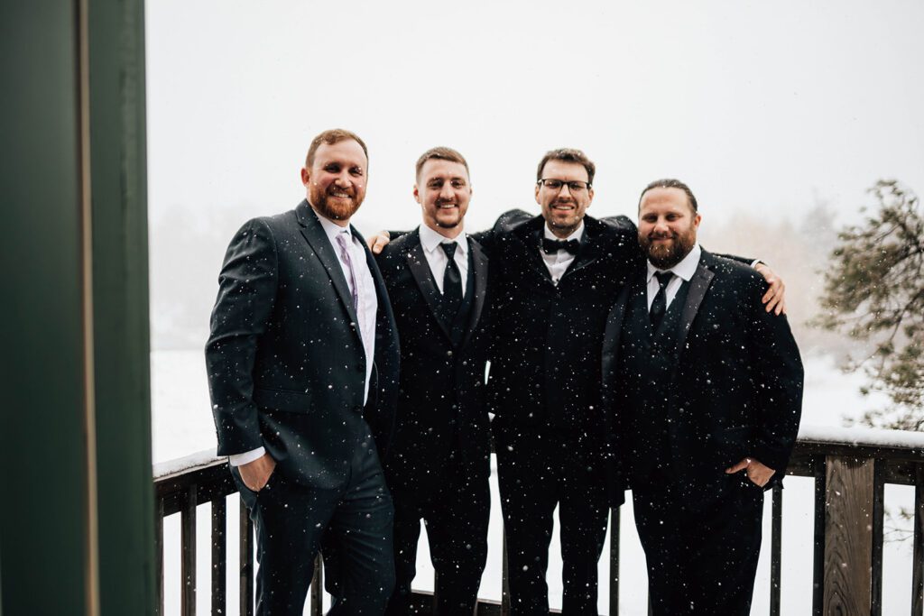 Groom and groomsmen smile together in suits on a snowy deck at The Arnold House, snowflakes falling around them.