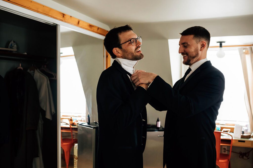 Groom laughs as a groomsman helps adjust his collar inside The Arnold House.