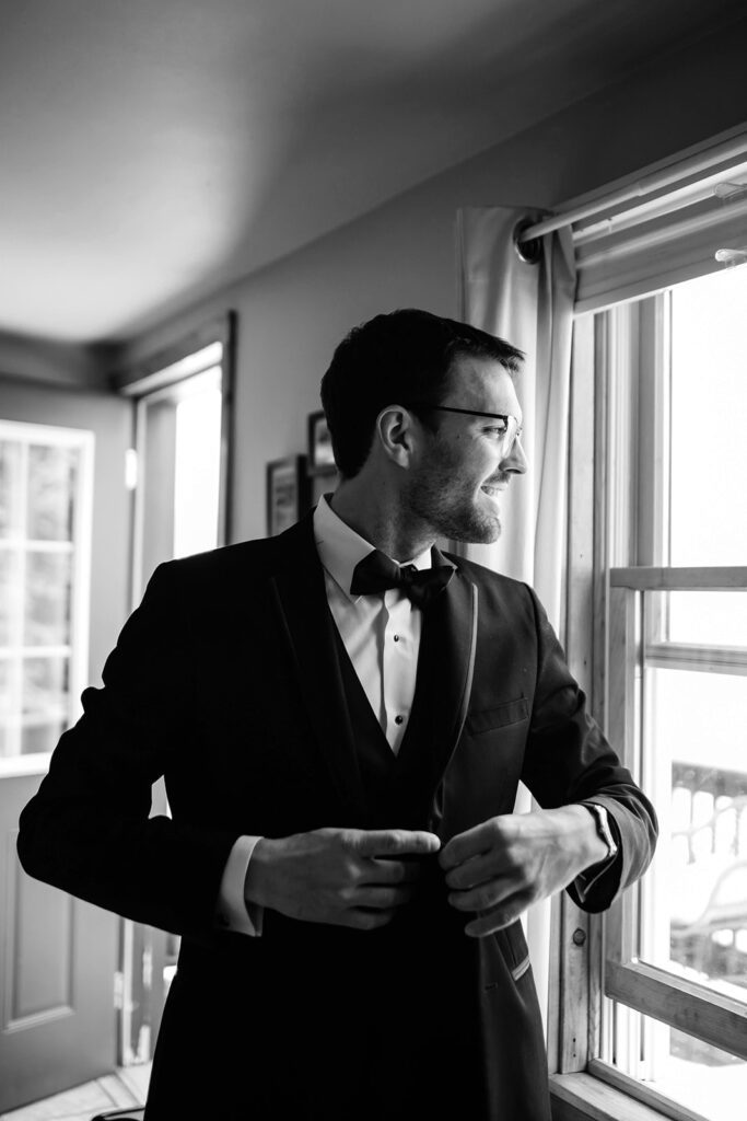 Groom smiles while fastening his suit jacket by a window.