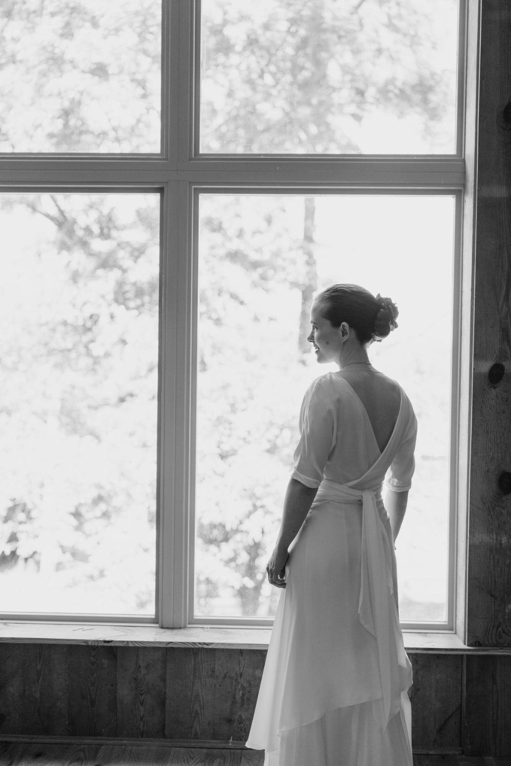 BLACK AND WHITE SHOT OF BRIDE STANDING BY A BRIGHT OPEN WINDOW