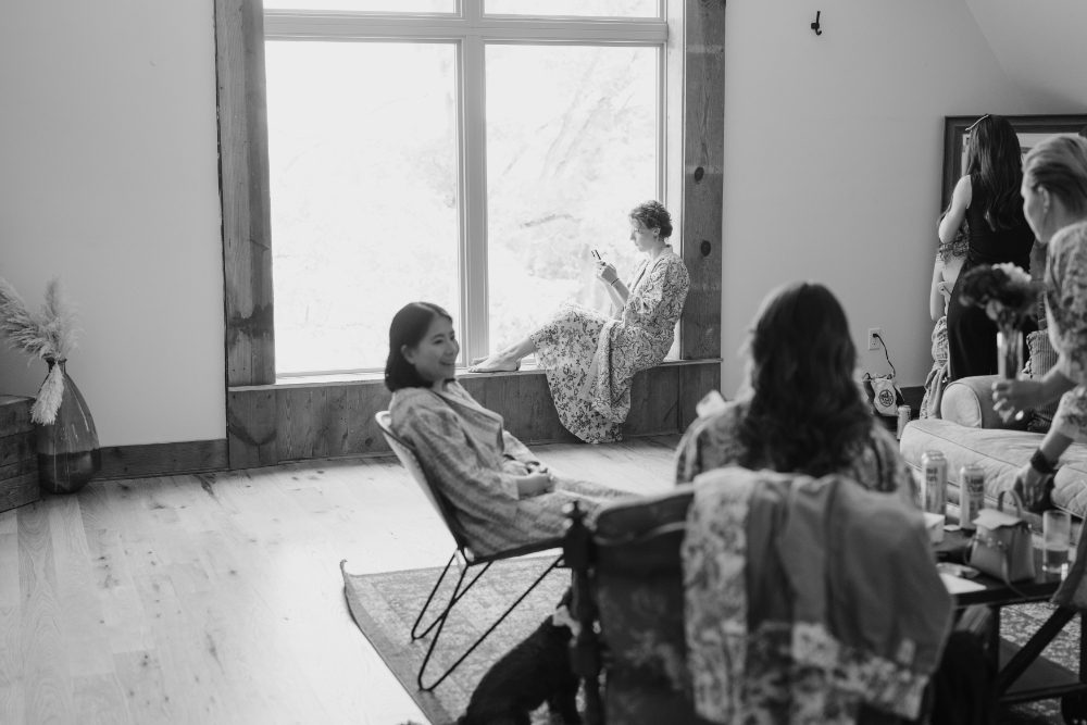 BLACK AND WHITE SHOT OF BRIDE AND BRIDESMAIDS GETTING READY IN A BRIGHT OPEN WINDOW