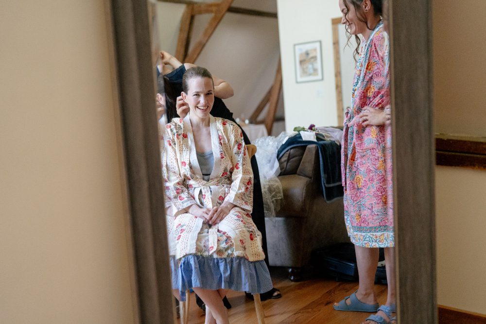 BRIDE SMILING INTO A MIRROW WHILE GETTING HER HAIR PINNED AND A BRIDESMAID SMILES BESIDE HER