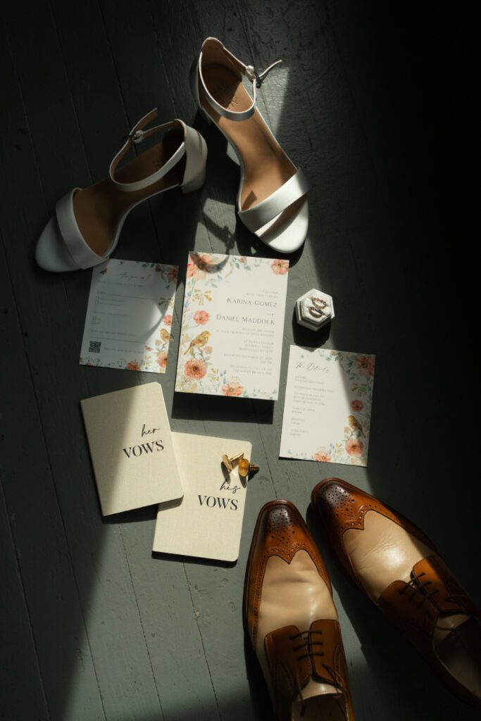 flat lay of whimsical floral wedding stationery, his and her vows, cuff links, wedding bands and bride and groom shoes.