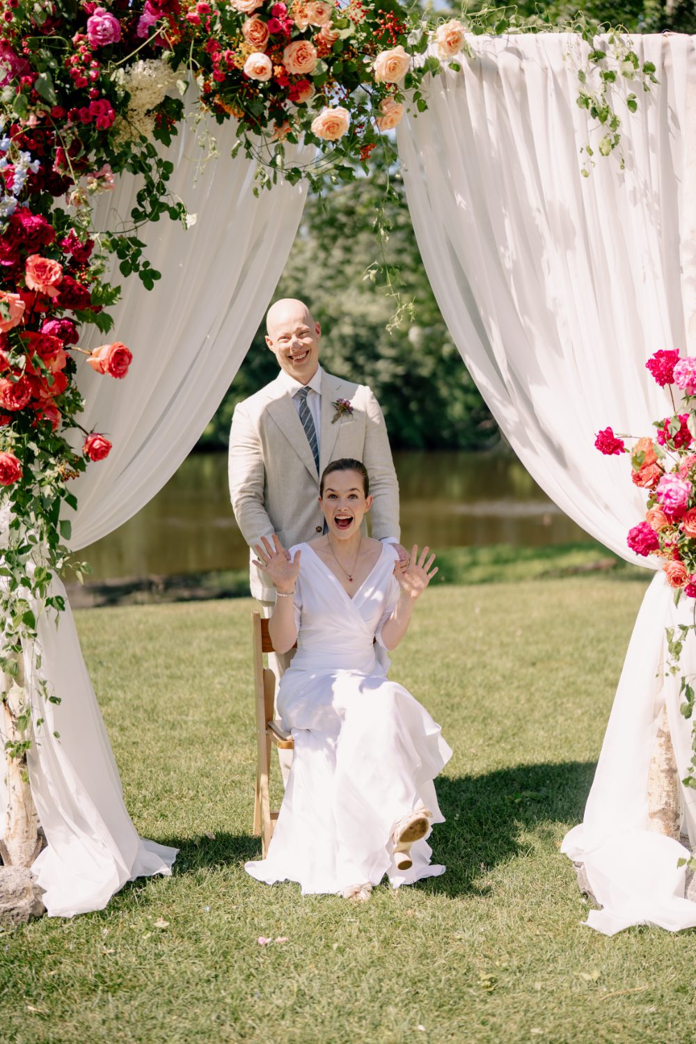 BRIDE SITS ON A WOOD CHAIR WITH GROOM BEHIND HER UNDERNEATH A WHITE DRAPED CEREMONY ARCH FILLED WITH CASCADING FLORALS