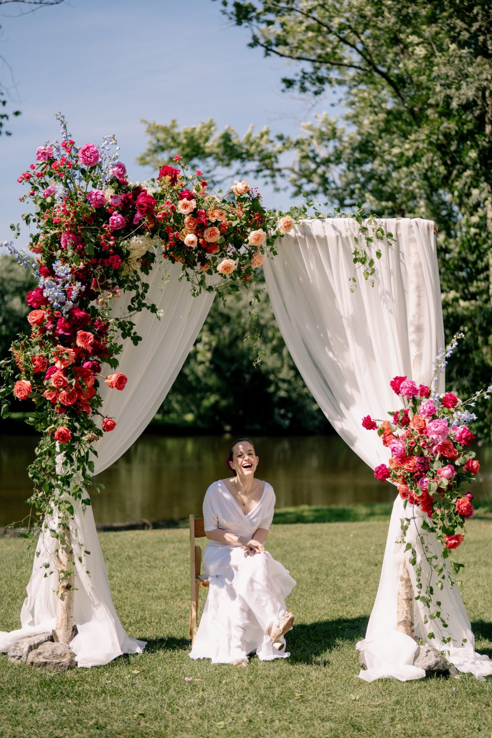 BRIDE SITS ON A WOOD CHAIR UNDERNEATH A WHITE DRAPED CEREMONY ARCH FILLED WITH CASCADING FLORALS