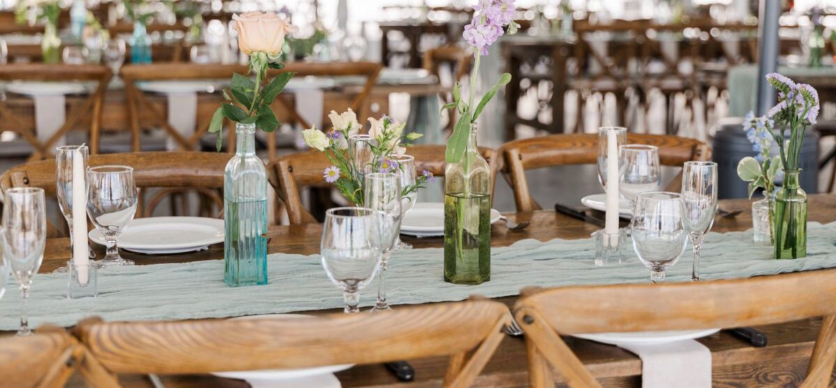 A long wooden reception table set with simple place settings, glassware, and soft pastel bud vases. The scene feels airy and inviting, with natural light filling a tented space and rows of wooden chairs creating a warm, relaxed atmosphere.