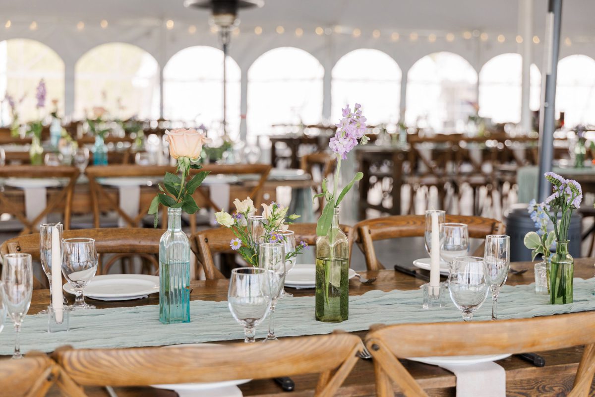A long wooden reception table set with simple place settings, glassware, and soft pastel bud vases. The scene feels airy and inviting, with natural light filling a tented space and rows of wooden chairs creating a warm, relaxed atmosphere.