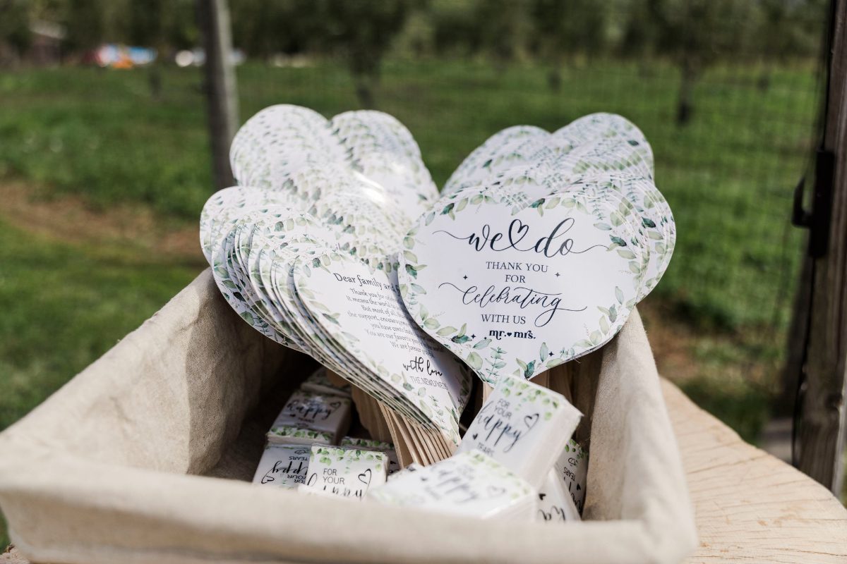 A basket filled with heart-shaped wedding fans decorated with greenery and soft script text. The fans sit outdoors on a sunny day, offering guests a sweet, personal welcome and a practical way to stay comfortable during the celebration. | how to plan a wedding step by step