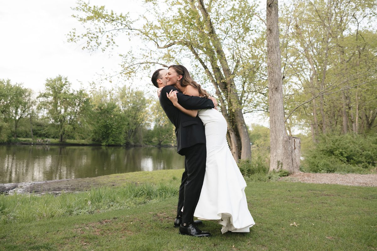 A newlywed couple embracing near a quiet pond surrounded by lush greenery. She wears a fitted white gown, and he wears a black suit. Their joyful hug captures a relaxed, heartfelt moment in a peaceful, natural setting.