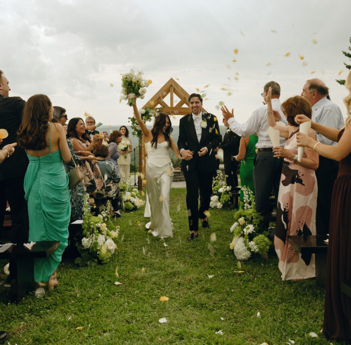 Newly married couple walk down an outdoor aisle as guests toss petals and cheer, bouquet raised in celebration beneath a wooden arch overlooking scenic countryside views.