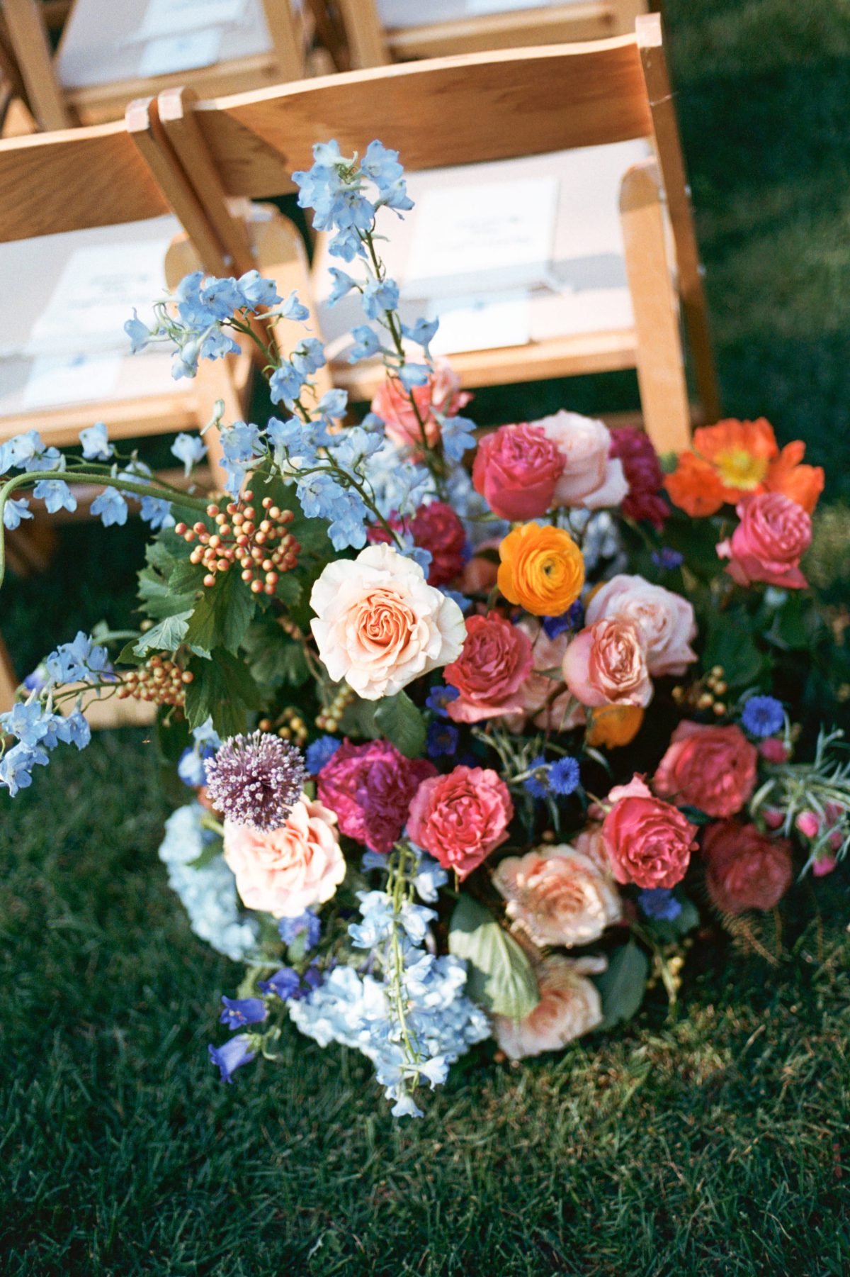 Colorful wedding ceremony flowers arranged beside wooden guest chairs, featuring roses and blue blooms, an example often shared in guides on how to save on a wedding using seasonal florals.