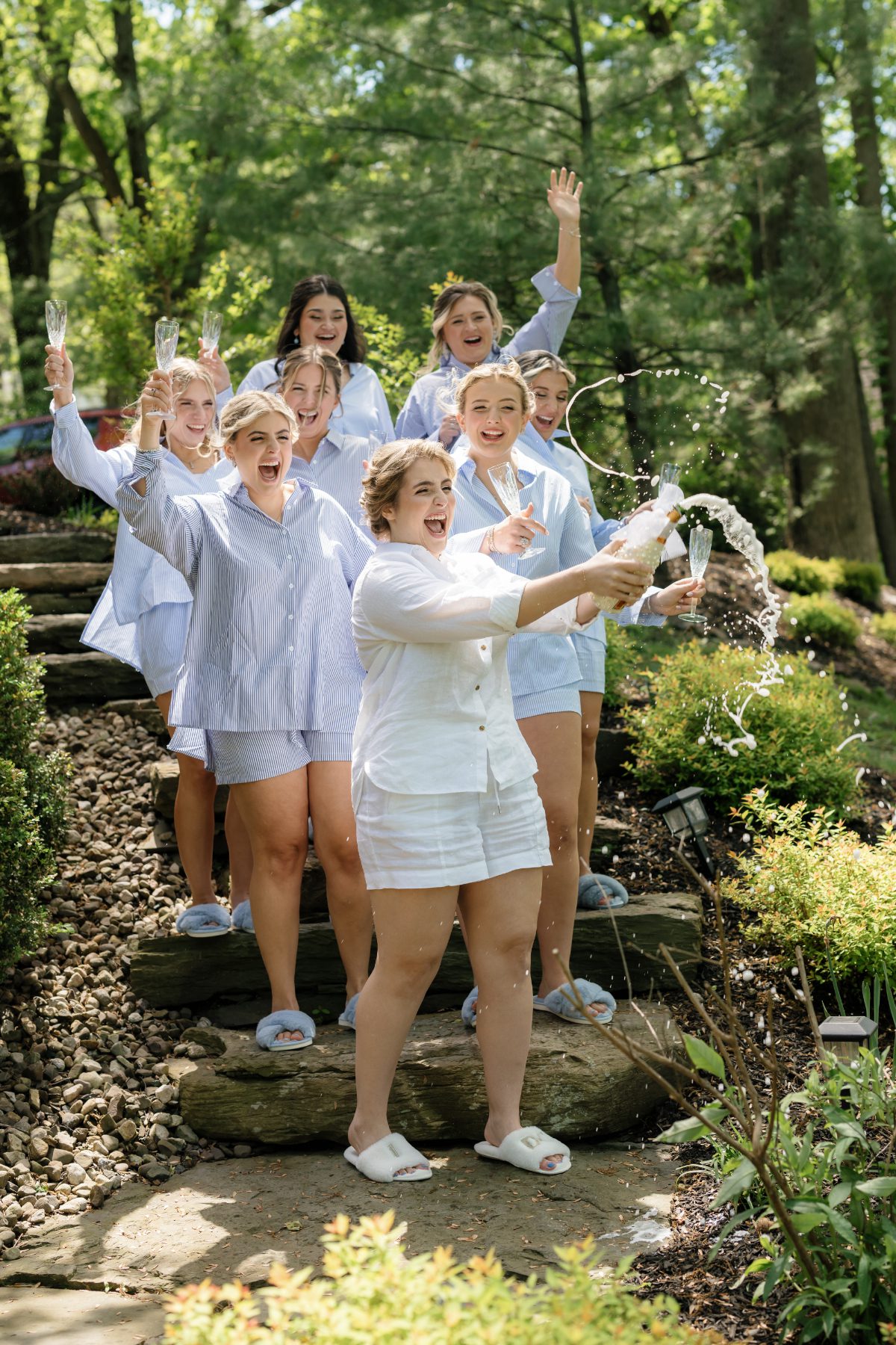 Wedding party in matching pajamas celebrate outdoors as one pops champagne on garden steps, laughing and raising glasses during joyful pre-ceremony festivities with friends under sunny trees.