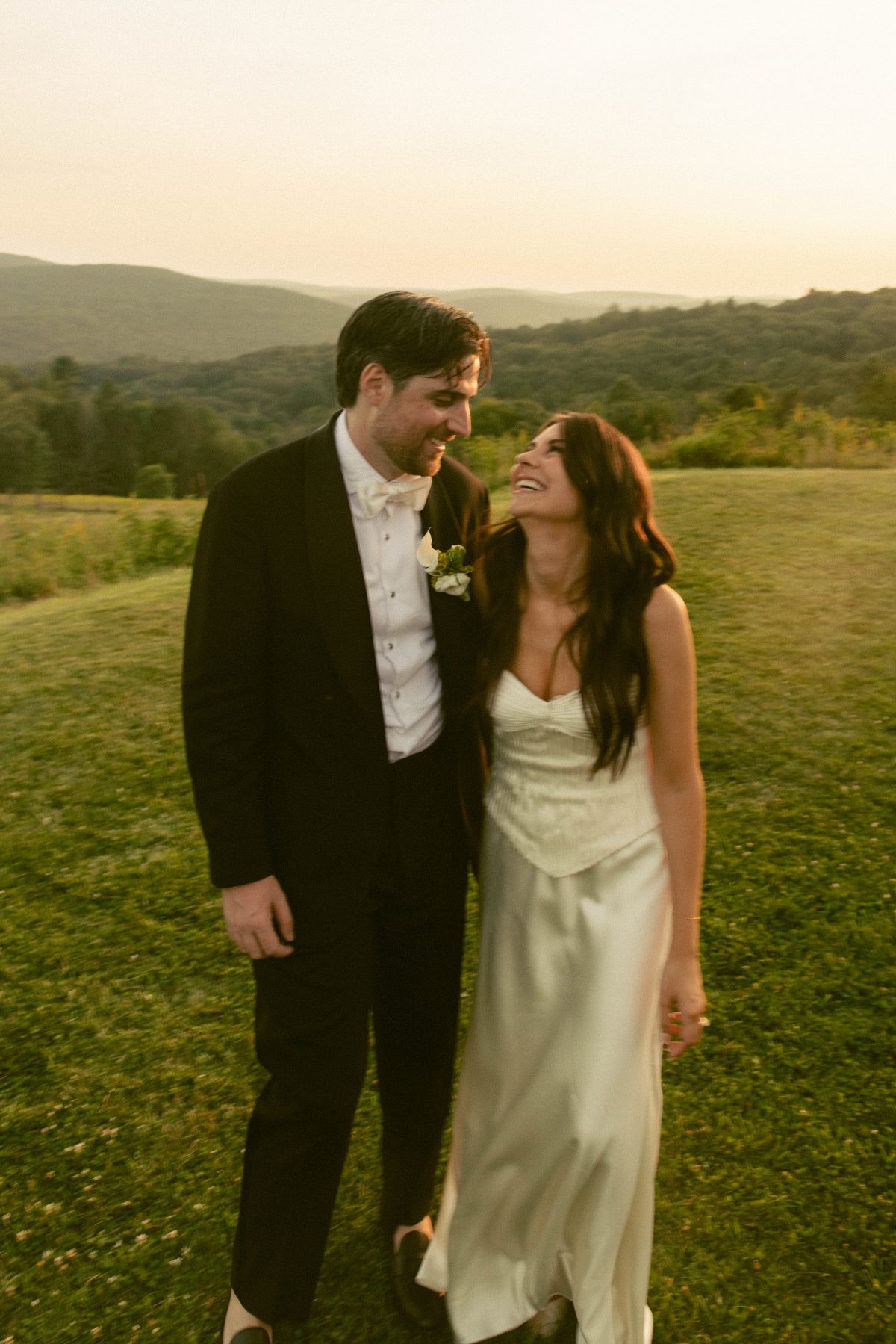 Bride and groom walk together at sunset across a grassy hill, smiling at each other as mountains glow behind them, capturing a relaxed, romantic moment after their outdoor wedding ceremony.