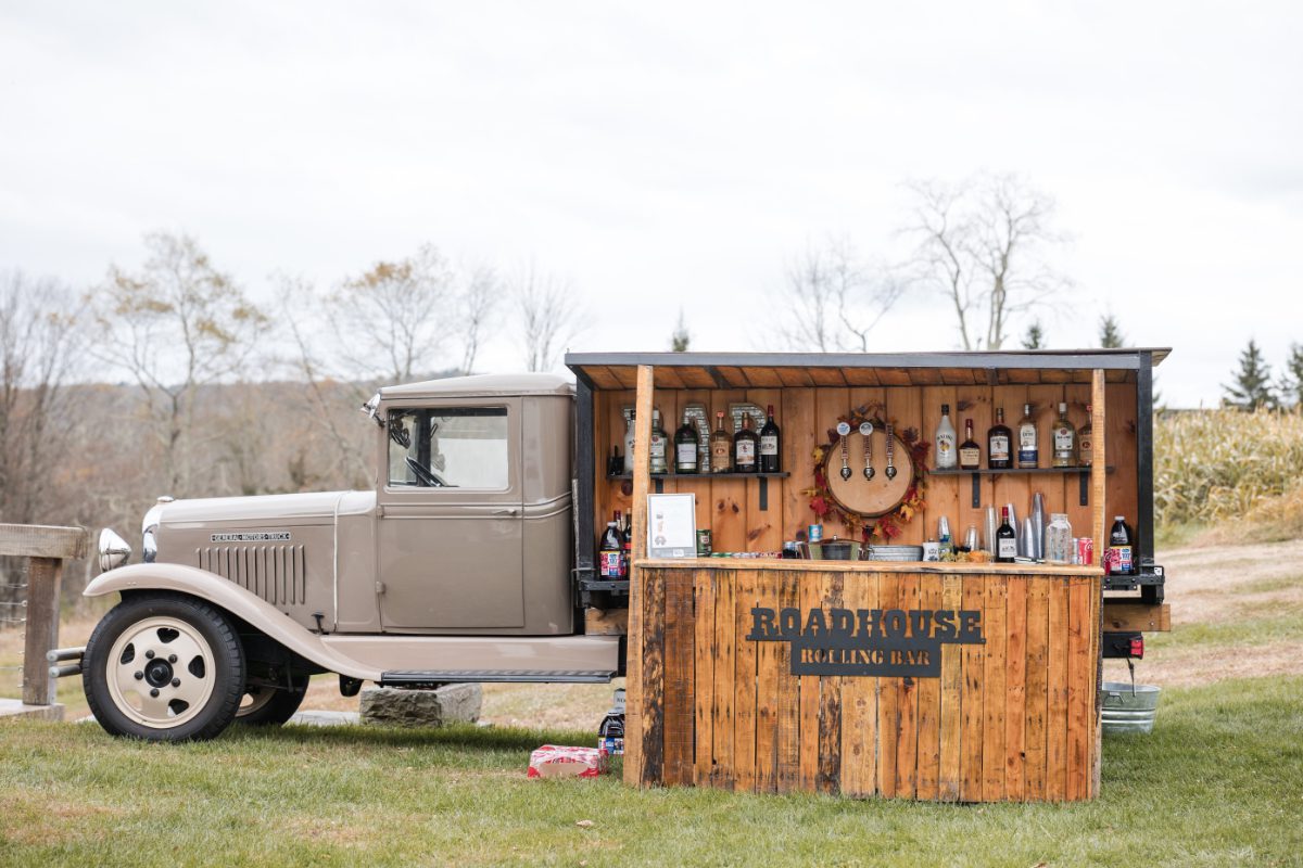 Vintage pickup transformed into a rustic mobile bar set on a grassy outdoor venue, stocked with bottles and taps, ready for guests at a countryside wedding reception or engagement celebration.