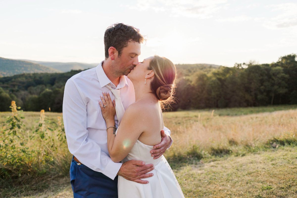 Newly married couple share a sunset kiss in a scenic field, embracing after their ceremony, with warm golden light and forested hills creating a romantic backdrop for wedding portraits.