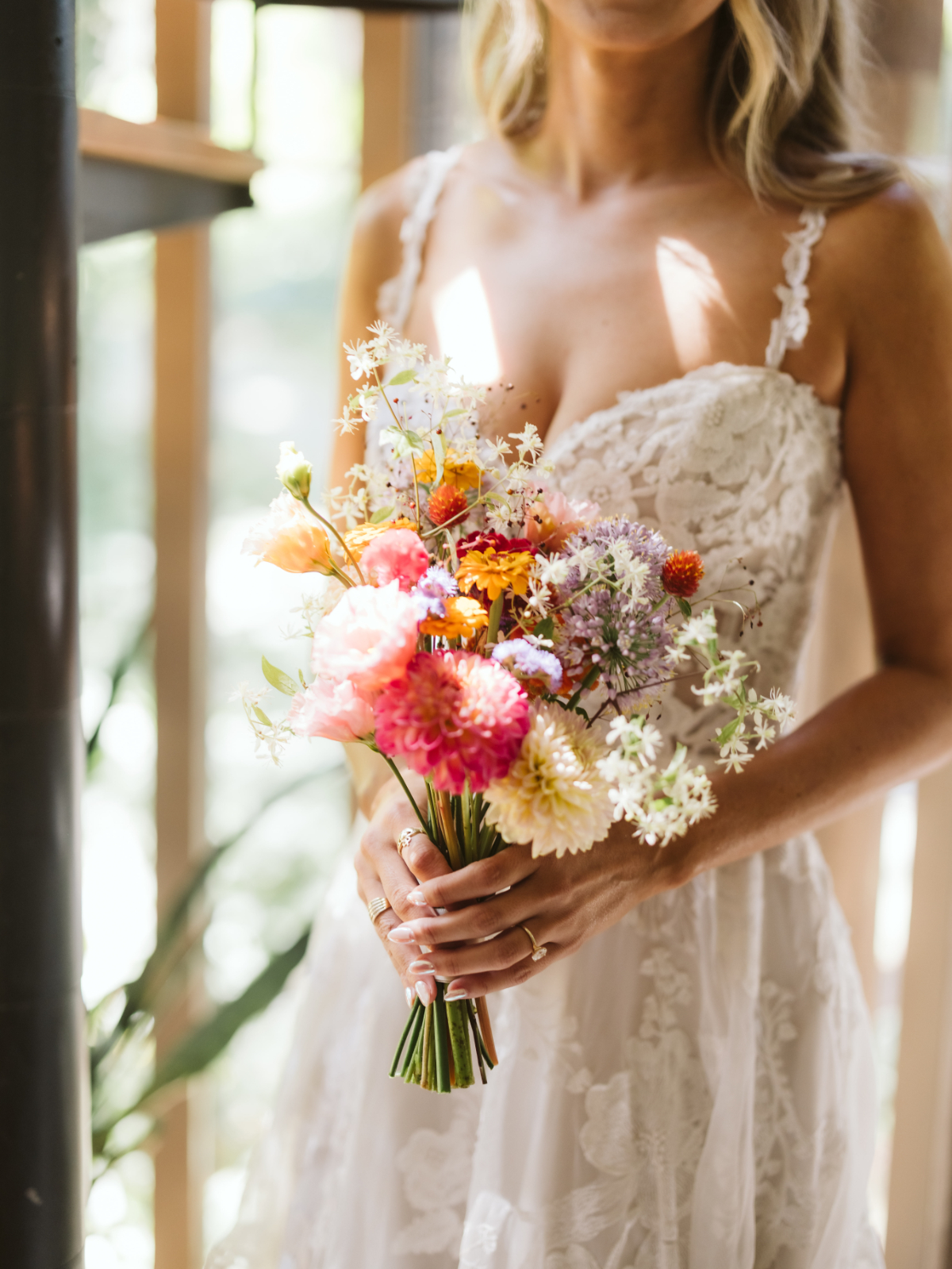 Bride in lace wedding gown holding a colorful wildflower bouquet indoors, sunlight highlighting floral details and textures, capturing a calm, intimate pre-ceremony portrait moment.
