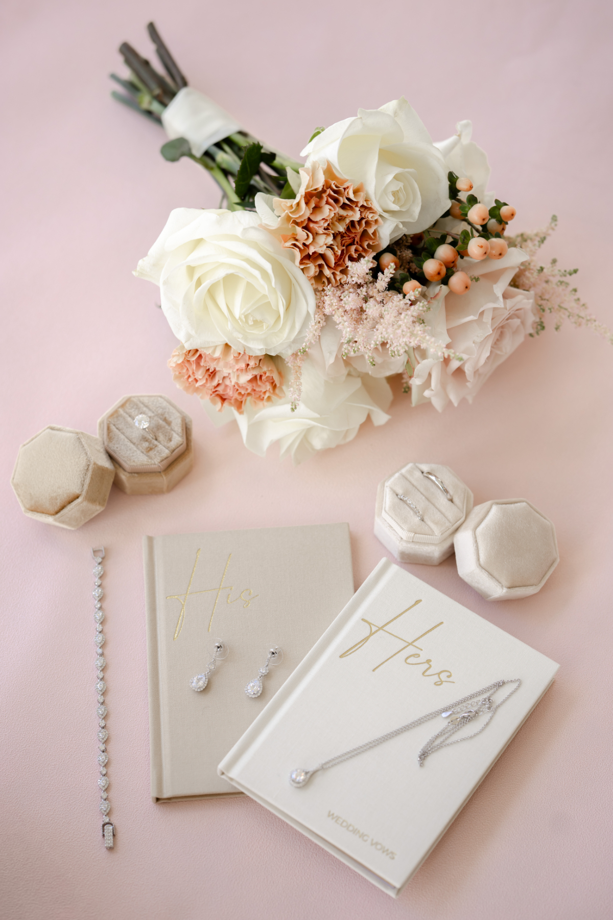 Wedding flat lay featuring bouquet, vow books, rings, bracelet, and earrings on soft pink background, highlighting elegant details and accessories prepared for the ceremony day.