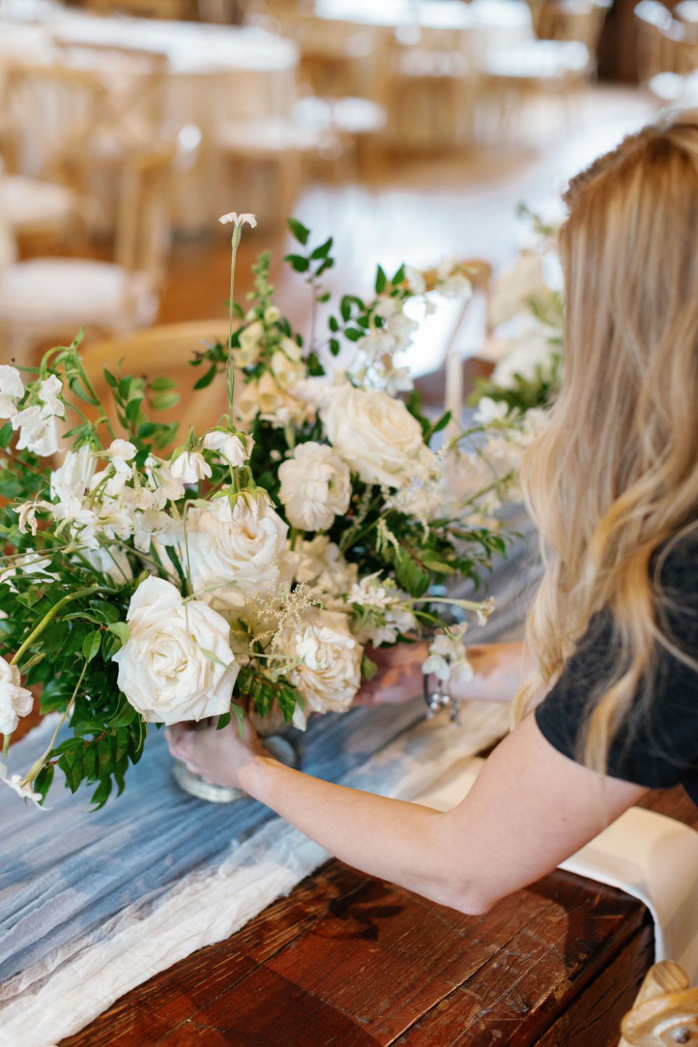 Close-up of a wedding planner placing a lush white and green floral arrangement on a wooden reception table with a soft blue runner, styling details for a Hudson Valley wedding weekend celebration.
