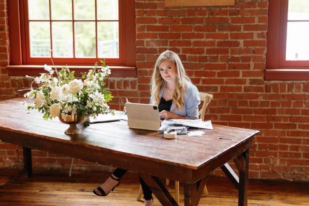 Wedding planner Kayce Drasher from KR Occasions sits at a rustic wooden table with her tablet, notes, and a lush white floral arrangement, preparing details for planning a wedding weekend in Hudson Valley.
