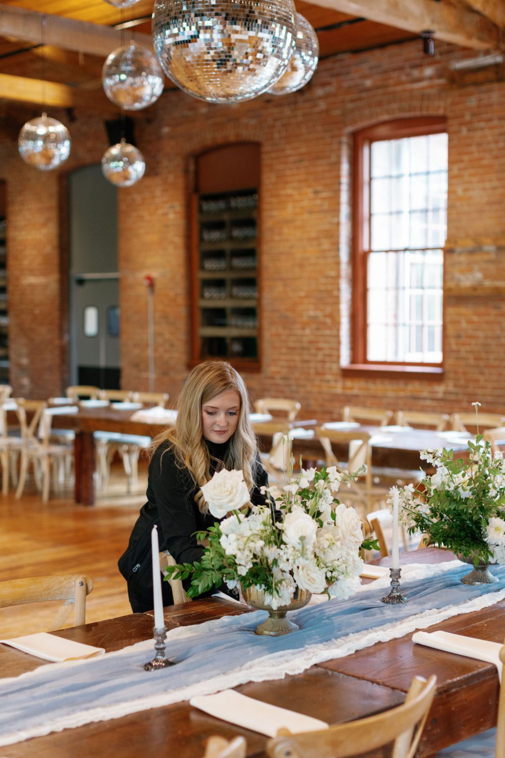 KR Occasions planner adjusts white roses and greenery on a farm table set with candles and a soft blue runner inside a brick-walled Hudson Valley reception space with disco ball accents overhead.
