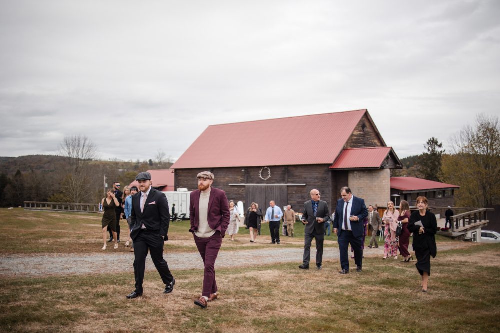 Wedding guests walk across the grounds of a rustic red-roofed barn venue in the Hudson Valley, arriving for the next event during a relaxed multi-day wedding weekend celebration.