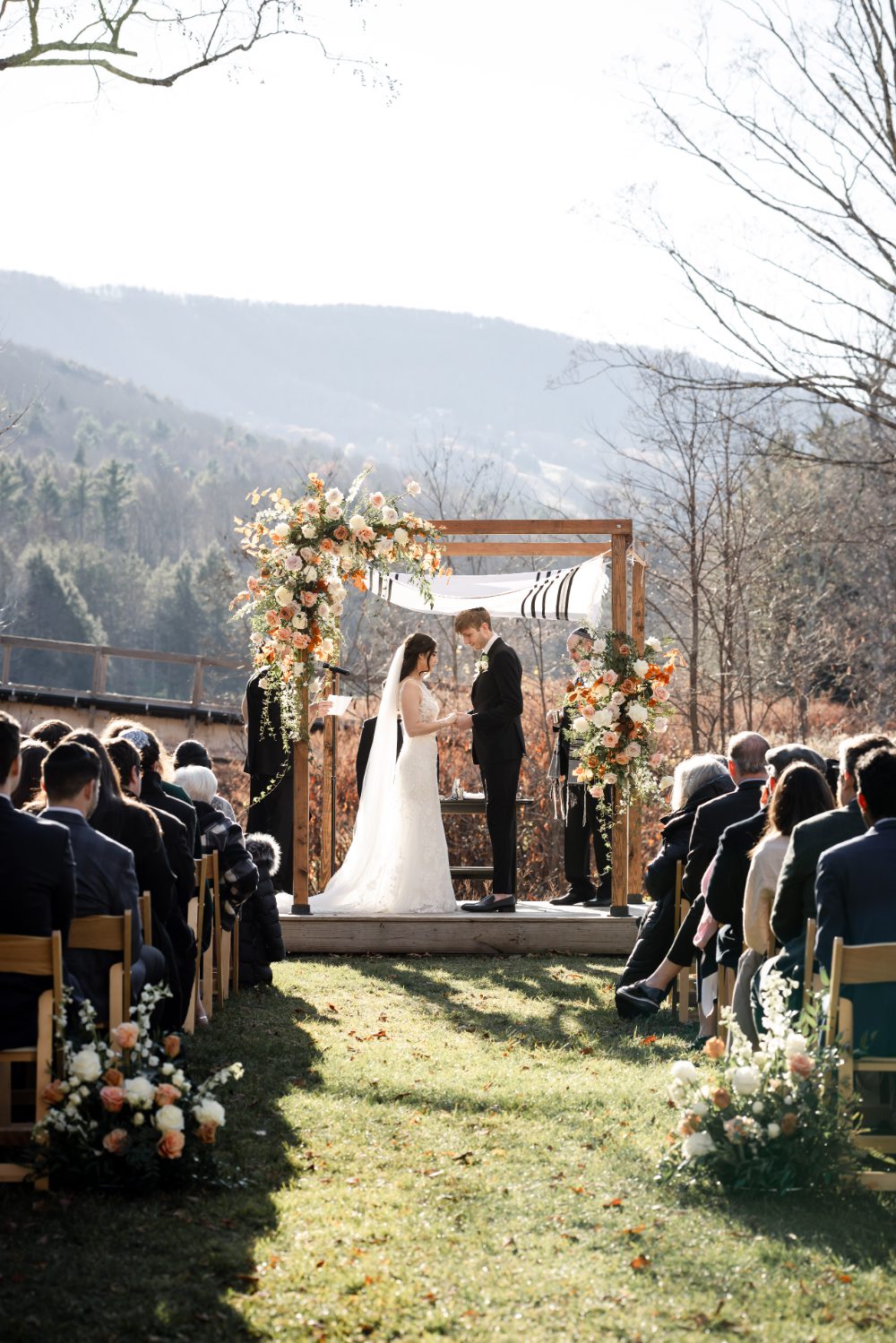 Couple exchanges vows under a wooden chuppah with peach and white floral arrangements as guests watch during an outdoor Hudson Valley ceremony with rolling mountains and late autumn light behind them.

